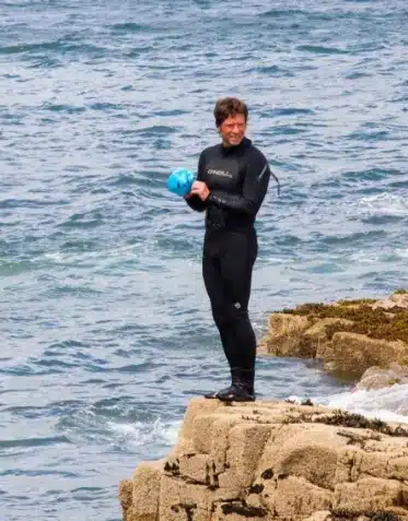 swim-coach-img-1 A person in a black wetsuit stands on a rocky outcrop by the ocean, holding a blue ball and smiling. Waves crash against the rocks beneath them, and the sea extends into the background.