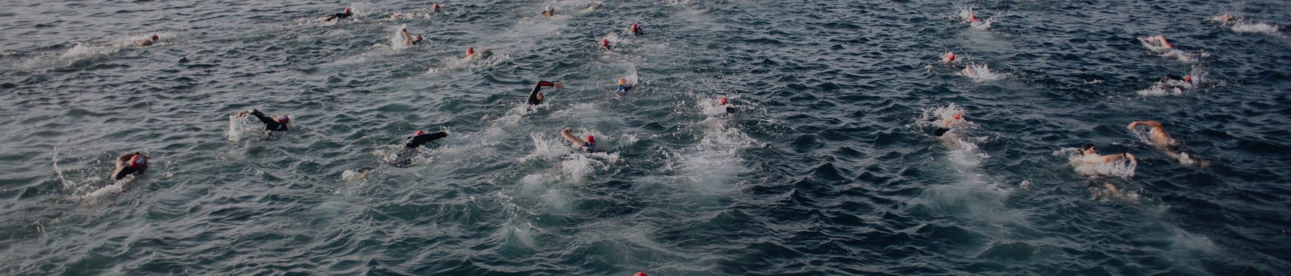 A group of swimmers with red swimming caps participating in an open water race, spread out across the choppy ocean surface. The swimmers create splashes as they move through the water. The daylight adds a blue tint to the scene.